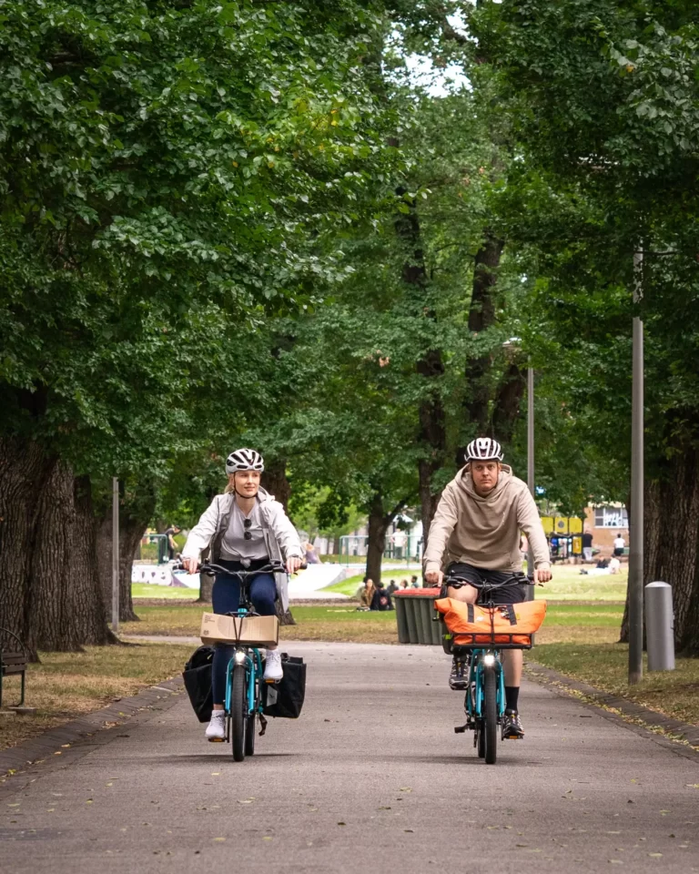 The image shows Kim and Nick enjoying a bike ride through a park in Melbourne on their Lug+Carrie Tern Quick Haul eBikes. The bikes have sturdy frames, rear cargo racks with panniers, and are equipped with electric pedal-assist systems. Kim is riding on the left-hand side of the image, wearing a helmet and casual clothing, while Nick is riding on the right-hand side, also wearing a helmet and carrying a backpack. They are both smiling and appear to be enjoying their ride through the park. The background shows a lush green landscape with trees, grass, and a paved path. The image conveys the joy and freedom of cycling, particularly on an eBike with pedal-assist technology, which makes cycling easier and more accessible. It also promotes the benefits of spending time outdoors, exercising, and exploring nature.