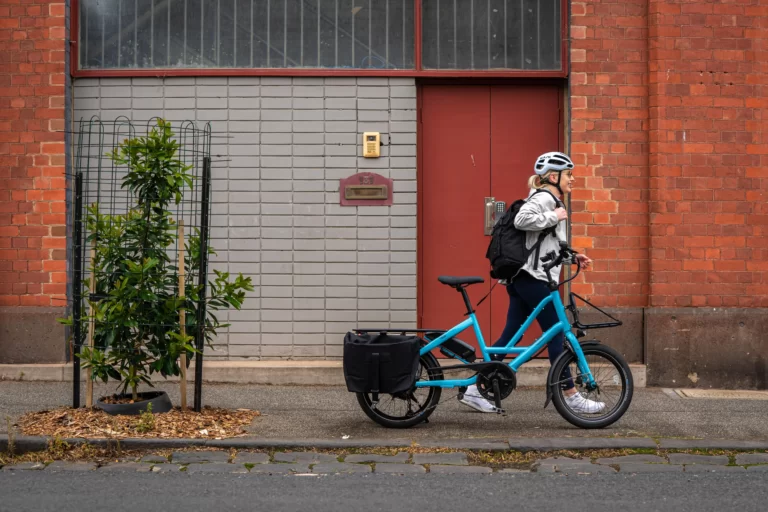 The image shows a woman walking past her parked Quick Haul bike from Lug+Carrie. The bike is parked on a sidewalk, and the woman is walking on the same sidewalk, beside the bike. The Quick Haul bike is designed for urban commuting and cargo-carrying capabilities, with a sturdy frame and a rear cargo rack with a basket. The woman appears to be using the bike for transportation, as she is carrying a backpack and is dressed in casual clothing. She is walking away from the bike, suggesting that she has just parked it and is continuing her journey on foot. The image conveys the convenience and ease of using the Quick Haul bike from Lug+Carrie for urban commuting and cargo transport, and the versatility of being able to park the bike and continue on foot.