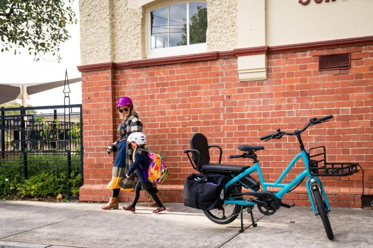 The image depicts a mother and daughter walking at a school after riding their Tern Quick Haul bike to school. The mother is holding the handlebars of the bike, which has a sturdy frame and a rear cargo rack equipped with a child seat. The daughter is walking beside the bike, and both mother and daughter are dressed in casual clothing and carrying backpacks. The image shows a school building in the background, suggesting that the mother and daughter are dropping off the daughter at school. The image conveys the convenience and practicality of using the Tern Quick Haul bike for family transportation, particularly for getting to and from school. It also highlights the bike's cargo-carrying capabilities and child-friendly features, such as the child seat.