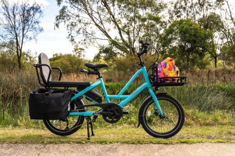 The image shows the Quick Haul eBike from Lug+Carrie with text overlay that reads "Affordable. Simple. Powerful. Everyday practicality to turn everyday chores into mini adventures." The bike has a sturdy frame, a rear cargo rack with panniers, and is equipped with an electric pedal-assist system. The background is a gradient of blue and green, suggesting a natural and outdoor environment. The text promotes the Quick Haul eBike as an affordable and simple solution for everyday practicality, such as running errands or commuting, that can be transformed into mini adventures. It emphasizes the bike's powerful electric pedal-assist system, which makes cycling easier and more accessible for people of all ages and fitness levels. The image conveys the practicality and versatility of the Quick Haul eBike, as well as the sense of freedom and adventure that cycling can provide.