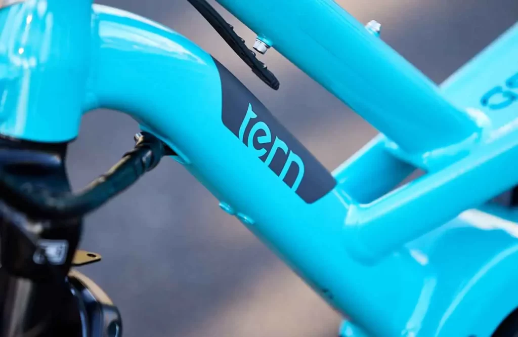 The image shows a close-up of a blue Tern ebike, designed for urban commuting and versatile cargo-carrying capabilities. The bike has a compact frame and is equipped with a powerful electric motor and battery, providing assistance while riding. The image showcases the bike's details, including the sleek blue frame, the Tern logo on the frame, and the electric motor and battery mounted on the downtube. The bike is equipped with several accessories, including fenders to protect the rider from splashes and a rear cargo rack to hold cargo or a child seat. The image conveys the quality and attention to detail in the Tern ebike's design, making it a practical and stylish option for urban commuters or families.