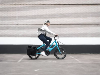 The image shows a mother and her daughter riding their blue Quick Haul eBike from Lug+Carrie on their way to school. The bike has a sturdy frame, a rear cargo rack with a basket, and is equipped with an electric pedal-assist system. The mother is riding on the left-hand side of the bike, wearing a helmet and casual clothing, while her daughter is riding on the right-hand side, also wearing a helmet and holding onto the rear cargo rack. They both appear to be enjoying their ride and are smiling. The background shows a suburban street with houses and trees in the distance, suggesting that they live in a residential area. The image conveys the convenience and practicality of using an eBike like the Quick Haul from Lug+Carrie for family transportation, particularly for school drop-offs and pick-ups. It also promotes the benefits of cycling for exercise and reducing carbon emissions.